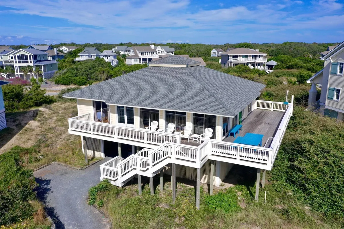The image showcases the exterior of a beautiful single-family beachfront home on Bald Head Island, North Carolina. The focal points include a spacious wraparound deck perfect for outdoor dining or lounging, expansive windows offering panoramic ocean views, lush greenery surrounding the property, and a welcoming staircase leading to the entrance.