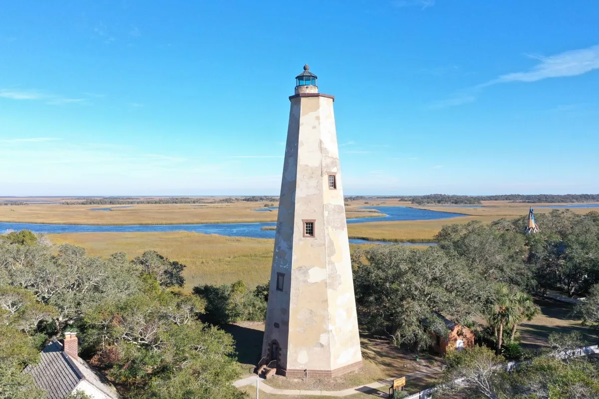 A classic lighthouse rises above surrounding marshlands and winding waterways, with the natural environment stretching into the distance. The scene highlights both the iconic structure and the diverse coastal ecosystem, including marshes that are ideal for birdwatching and observing other wildlife.