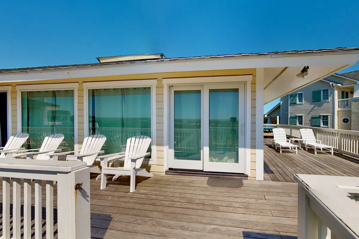 An inviting wooden deck with adirondack chairs facing out towards the ocean. Large glass doors connect the deck to the interior, reinforcing the idea of seamless indoor-outdoor flow. The tranquil atmosphere is accentuated by the possibility of relaxing, socializing, or enjoying sunset views.