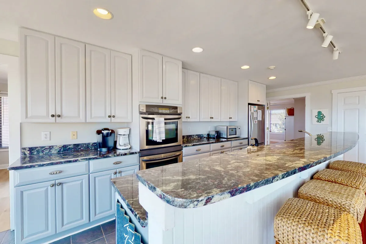 A close-up view of the modern kitchen area, featuring a granite countertop, central island, white cabinetry, and stainless steel appliances. The arrangement is ideal for cooking together, entertaining, or enjoying a morning coffee while taking in the fresh, bright atmosphere.