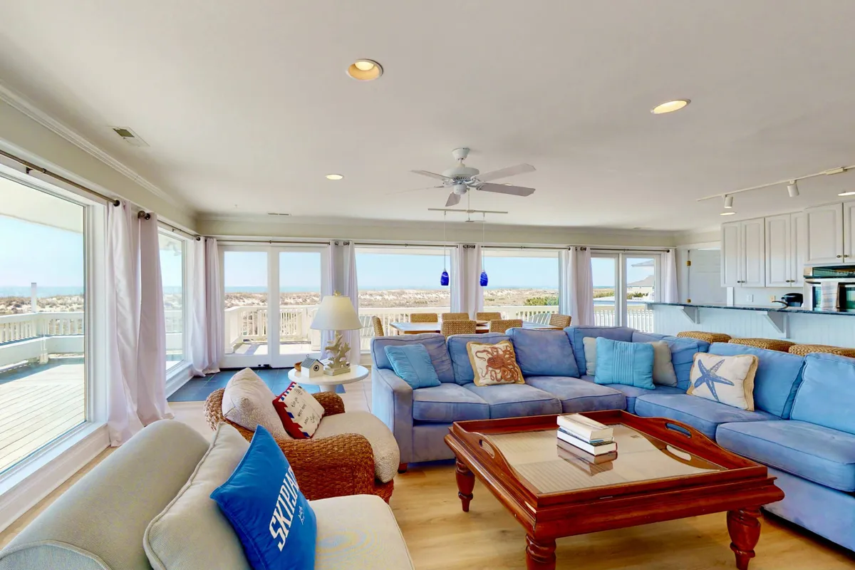 A spacious living room with a large sectional sofa faces a wall of windows overlooking the ocean. The arrangement encourages both relaxation and group activities like movie nights or games, with ample plush seating and a coffee table at the center.