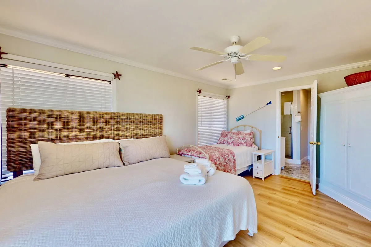 A tranquil bedroom featuring a wicker headboard, crisp bed linens, and neatly folded towels on the bed. A bedside table adds practicality, while soft lighting and neutral tones create a peaceful atmosphere ideal for unwinding after a day out.
