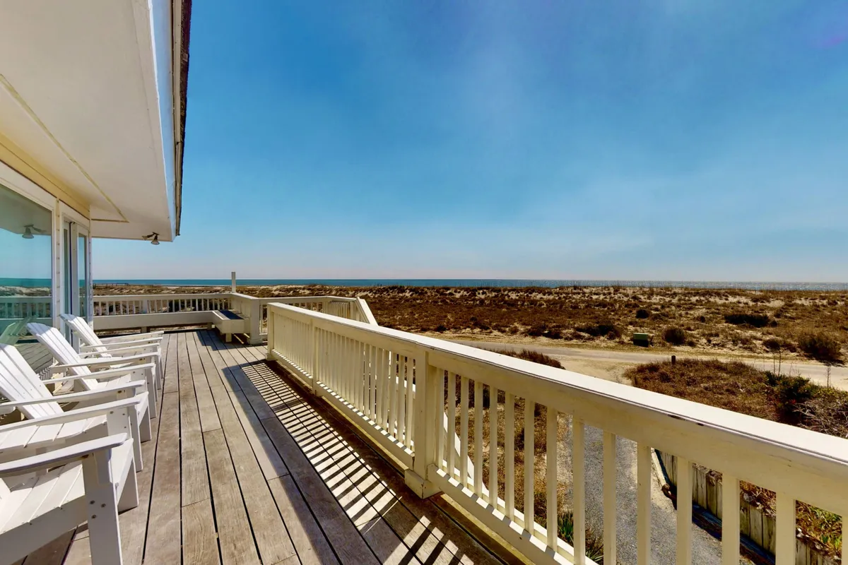 A view from the deck itself, highlighting the classic adirondack chairs positioned along the railing with a striking ocean view beyond. The image captures a tranquil sunset mood, emphasizing relaxation and the prime vantage point for watching the water or spotting dolphins.
