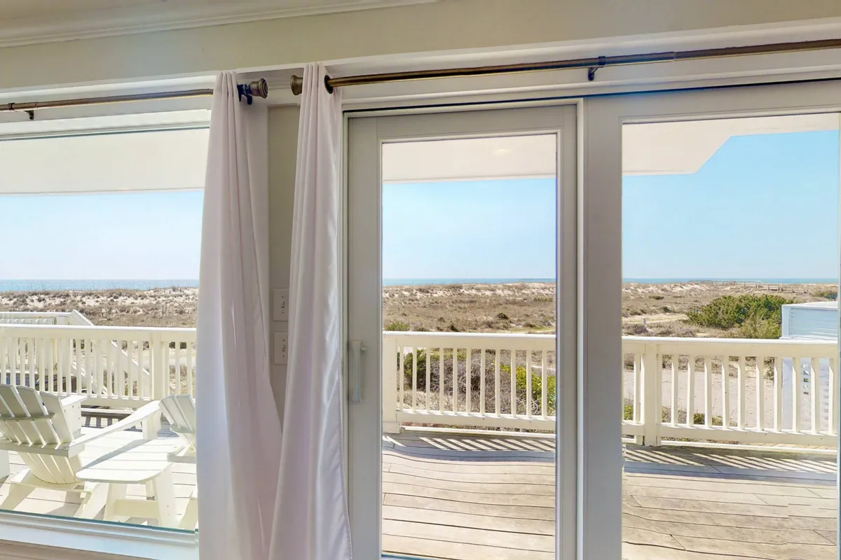 This image features large sliding glass doors with white curtains opening onto an outdoor deck, where chairs are visible, and the expansive ocean view beckons. The space blends the interior and exterior, inviting the outdoors in and highlighting a seamless transition to the wrap-around deck.