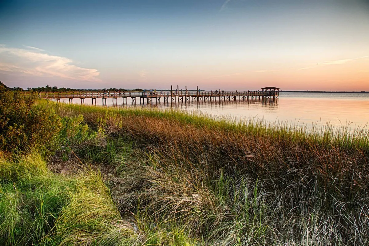 A tranquil wooden pier stretches out over calm water, surrounded by swaying tall grasses and framed by a stunning sunset sky. The scene exudes peace, ideal for fishing, picnicking, or capturing memorable sunset photographs.
