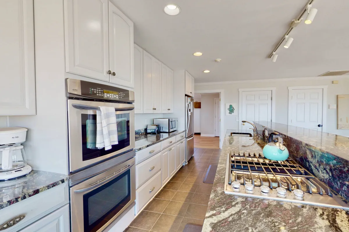 A bright, functional kitchen with granite countertops, stainless steel appliances, a kitchen island with seating, and a pop of color from a blue kettle. The space is open, practical, and ideal for prepping beach-day snacks or refueling after adventures.