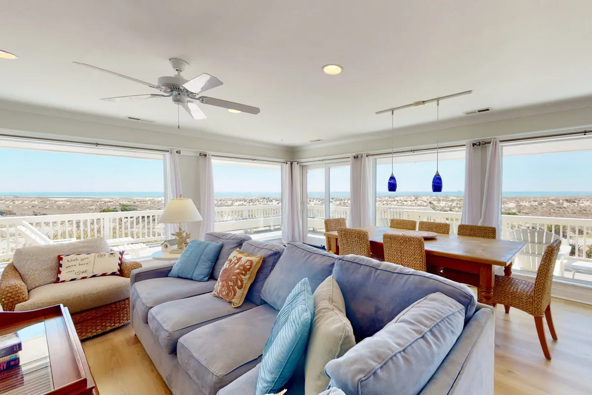 This image showcases a bright and welcoming living area featuring a comfortable blue sofa, a dining table with wicker chairs, and expansive windows that offer stunning ocean views. The setup emphasizes a calm, social, and airy atmosphere perfectly suited for gatherings or quiet relaxation, with the ocean as a constant backdrop.