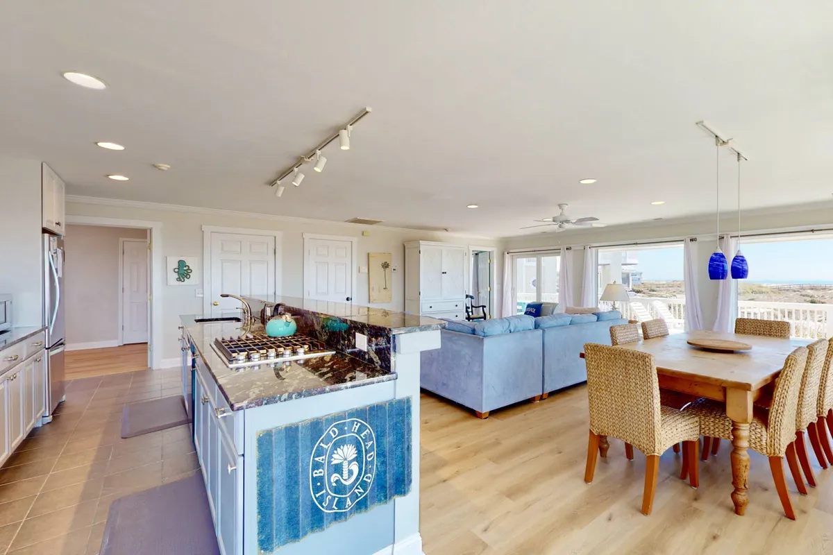 The image captures the integrated layout of the kitchen, dining, and living areas. Large windows provide abundant natural light and sweeping water views. The kitchen features a central island and modern finishes, inviting both meal preparation and social interaction in a cozy, welcoming setting.