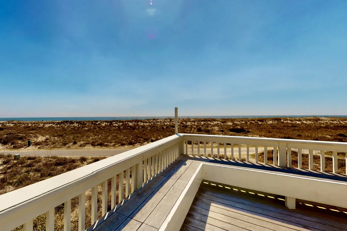 A sunlit deck with a wooden railing overlooks an expansive ocean view. The setting is peaceful, with the soft glow of either sunset or sunrise casting light across the water, creating a serene atmosphere perfect for unwinding or enjoying a quiet moment with a drink in hand.