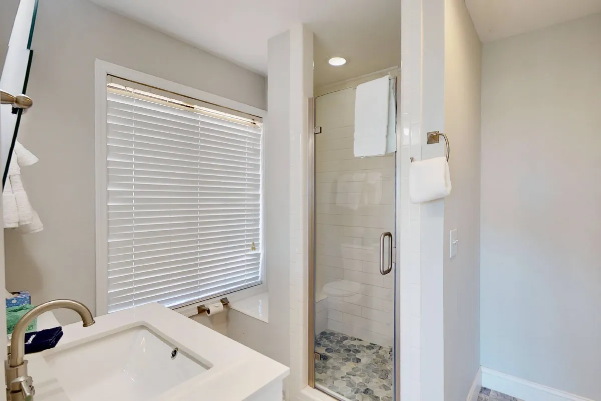 The image shows a modern bathroom with a sleek sink, glass shower enclosure, and ample natural light streaming through a window, creating a serene and fresh environment for guests to refresh and reset.