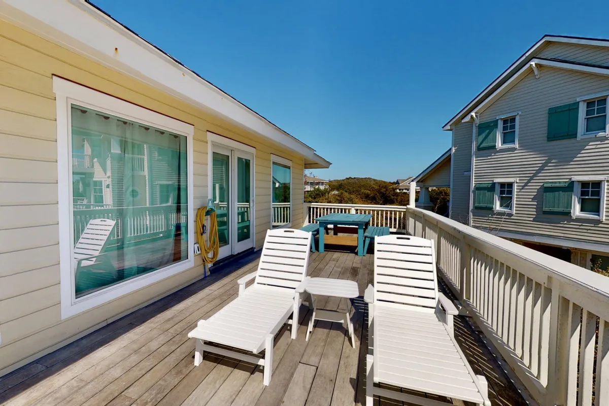 A ground-level exterior view showing lounge chairs and a table set up on a shaded patio beneath the deck, with windows above. This spot is perfect for casual outdoor dining, picnics, or prepping for beach days in a protected area.