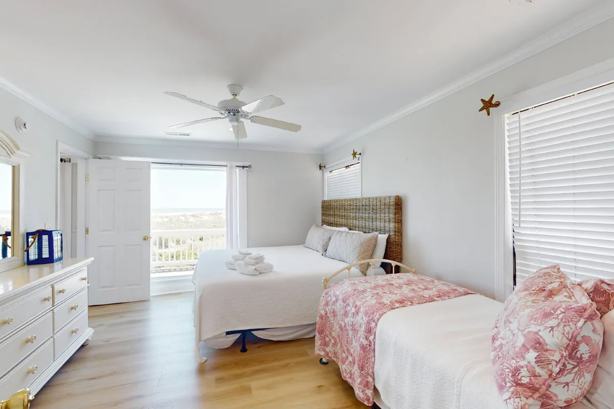 A tranquil bedroom with a bed featuring white linens and decorative pillows, a dresser with a mirror, and large windows letting in abundant natural light. The space feels cozy, perfect for relaxing and enjoying a quiet moment.