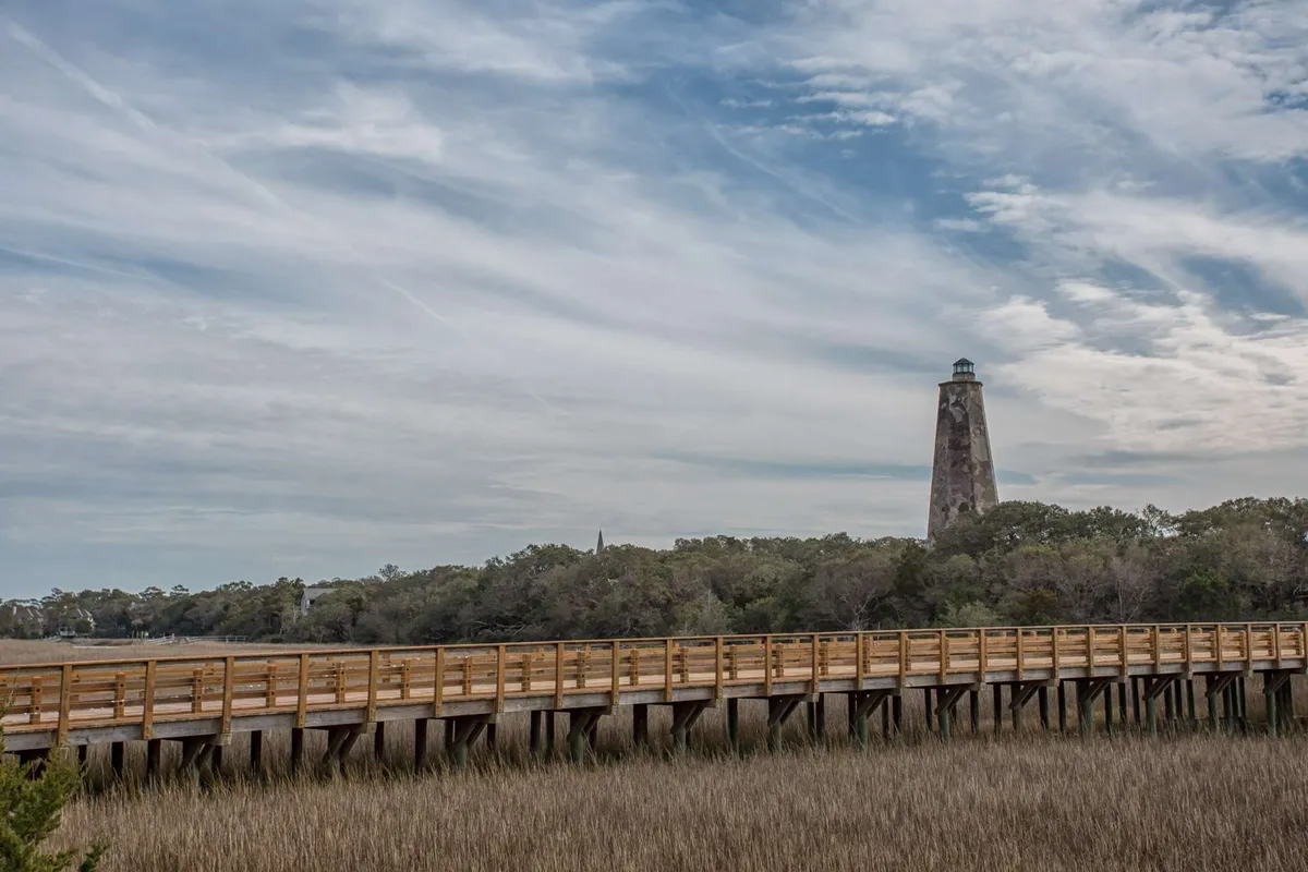 This image features Old Baldy lighthouse standing tall beside a wooden boardwalk that winds through lush marshland. The serene landscape is accentuated by soft natural light, and the scene invites exploration and discovery. The lighthouse dominates the composition, serving as a clear focal point and historical anchor.