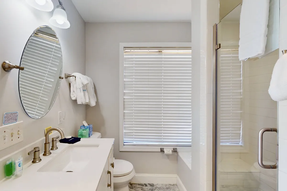 A modern bathroom featuring an oval mirror, double sink, shower enclosure, and a window with blinds. The design is fresh and spa-like, emphasizing privacy and the convenience of dedicated bathrooms for each suite.